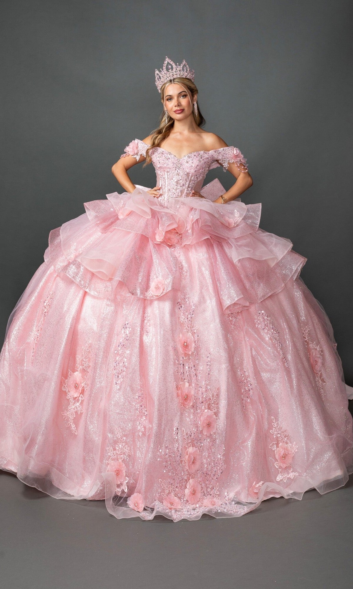 A young woman wears the Quinceanera Dress 5132 by Dancing Queen in pink, featuring a corset bodice and matching crown, as she smiles at the camera against a gray background.